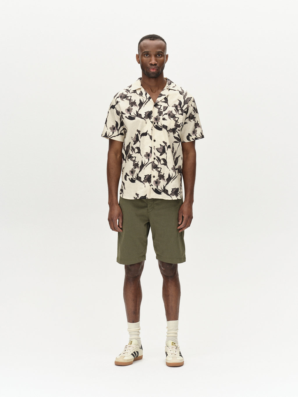 A man stands against a white background, wearing a short-sleeve shirt with a black and white abstract pattern, Gabbas Jet K3280 Dale Shorts in deep lichen green, and white sneakers with black stripes. Exuding modern style and all-day comfort, he faces forward in relaxed elegance.