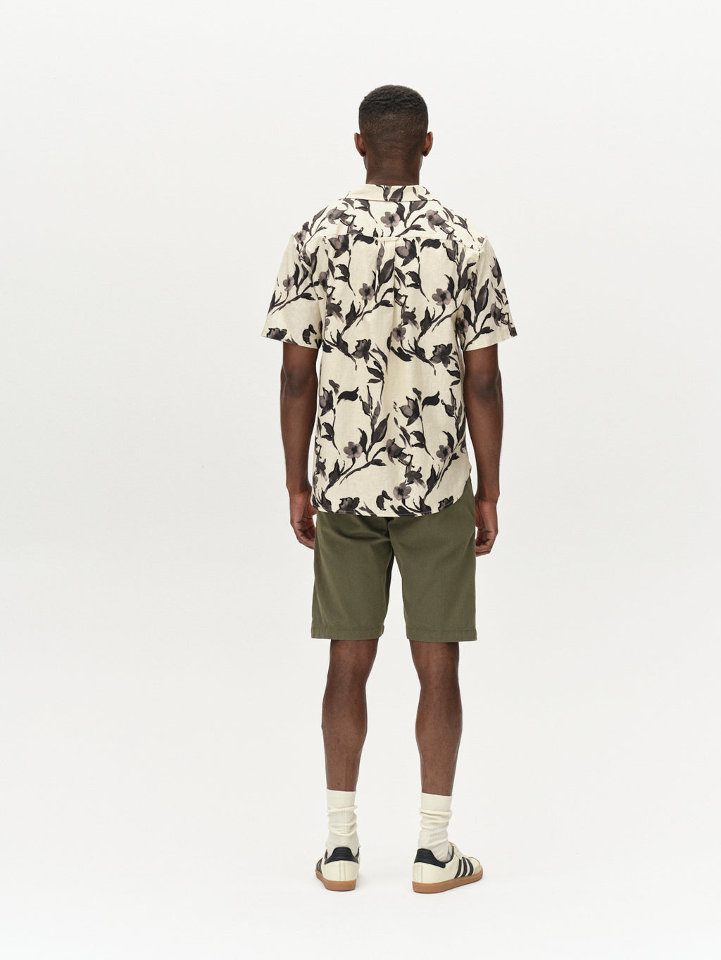 A person with short hair faces away, modeling a floral short-sleeve shirt paired with Gabbas Jet K3280 Dale Shorts in Deep Lichen Green. The ensemble, featuring white socks and striped shoes, radiates comfort and versatility against the plain backdrop.