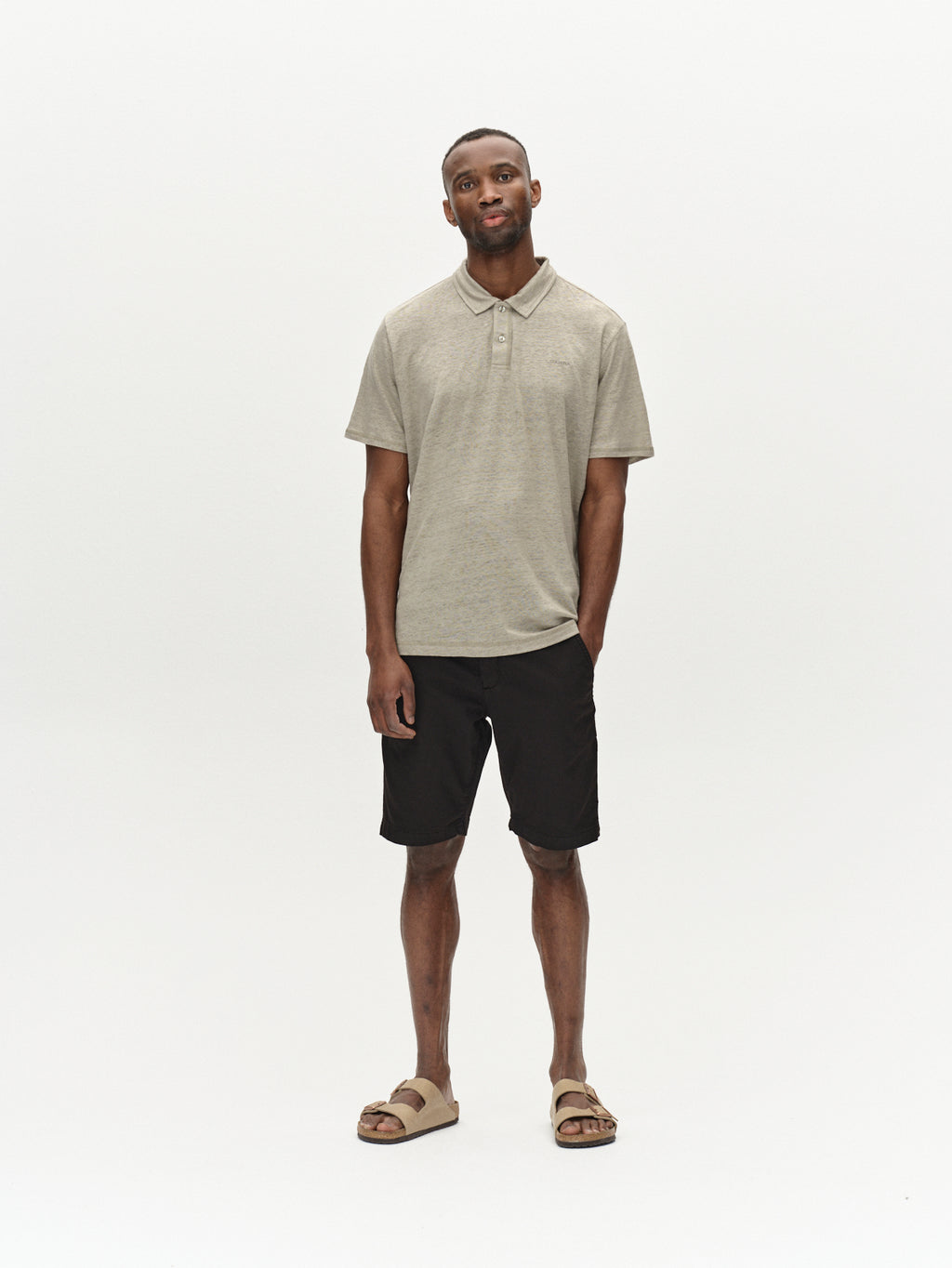 A person stands against a plain background wearing a light gray polo, Jet K3280 Dale Shorts in black by Gabba, and beige sandals. They exude effortless style with a relaxed posture and slightly tilted head.
