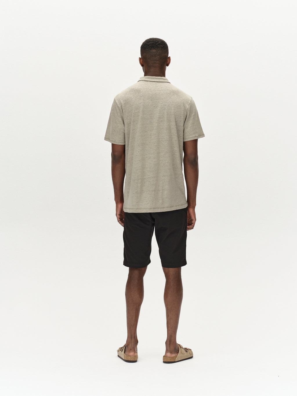 A man faces away from the camera, effortlessly styled in a beige polo shirt, Jet K3280 Dale Shorts - Black by Gabba, and sandals. Set against a plain white background, his look exudes modern design and all-day comfort.