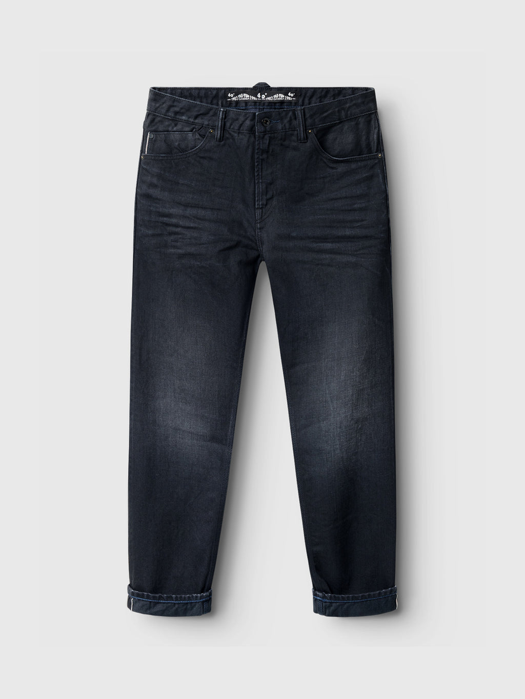 The Carl Selvedge 40th Anniversary jeans by Gabba feature a classic five-pocket design and button closure in dark blue-black denim with rolled-up cuffs, set against a light gray background for a durable and stylish look, displayed flat in a frontal view.