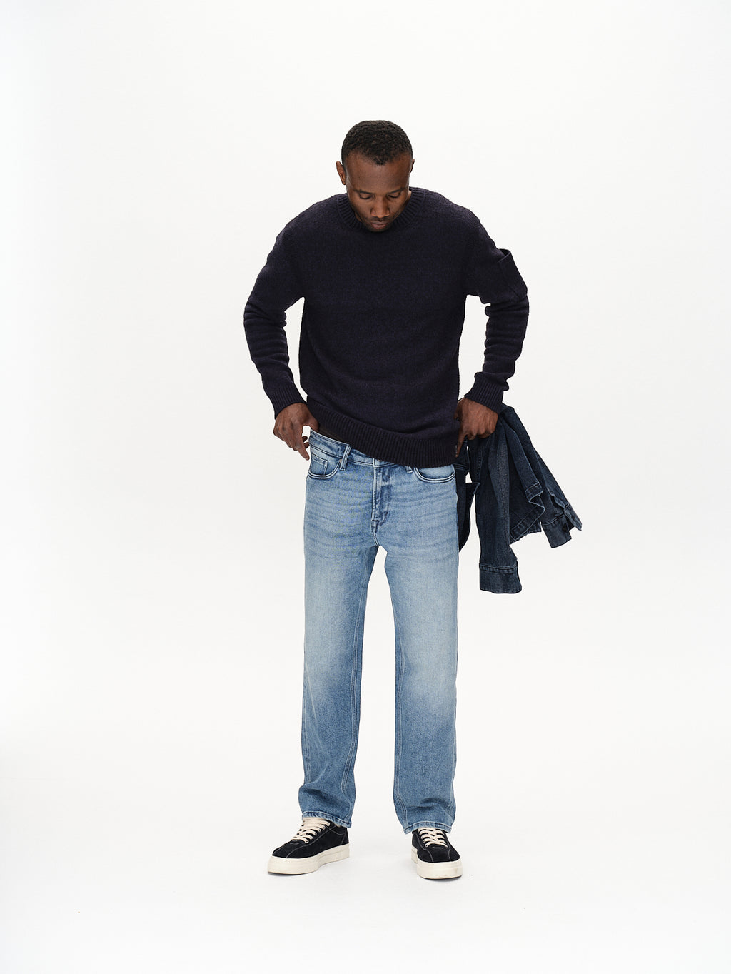 A person stands against a plain background, looking down. They wear the Luca Ls - Navy sweater from Gabba with light blue jeans and sneakers, holding a denim jacket in their left hand for a laid-back touch.