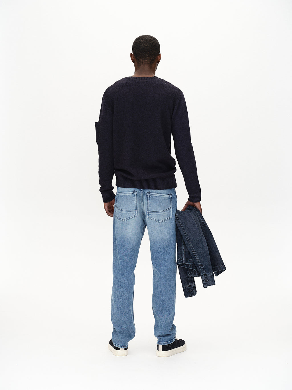 A person stands with their back to the camera wearing the Gabba Luca Ls - Navy sweater paired with light blue jeans and black shoes, holding a denim jacket in one hand against a plain white background.