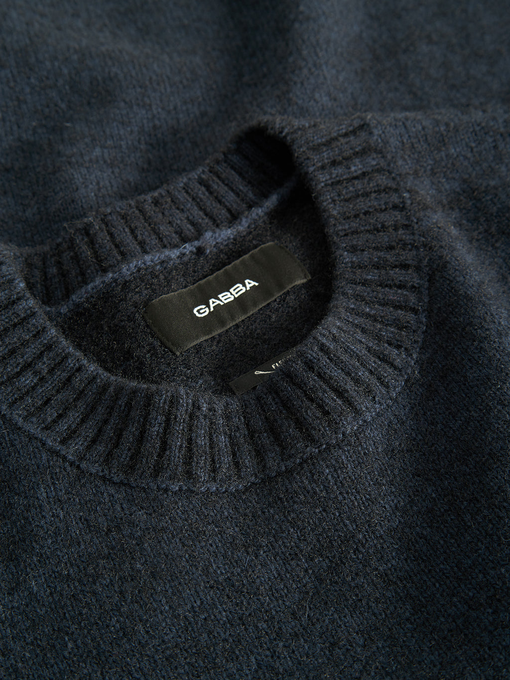 The Luca Ls - Navy from Gabba is a dark navy blue sweater featuring plaited knitting and a ribbed collar. A black tag with white Gabba text is near the neckline. Made from mixed regular wool, the fabric feels soft and slightly fuzzy.