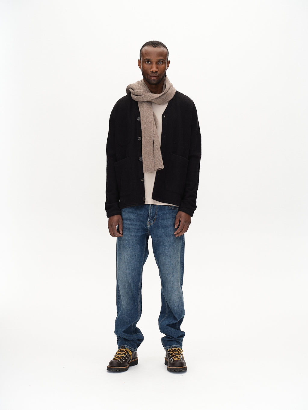 A person faces forward against a white background, wearing Gabbas Mattia Ls - Meteorite cardigan with flatlock stitching, a beige scarf, light beige shirt, blue jeans, and black boots with yellow laces. Their hands rest by their sides and they have a neutral expression.
