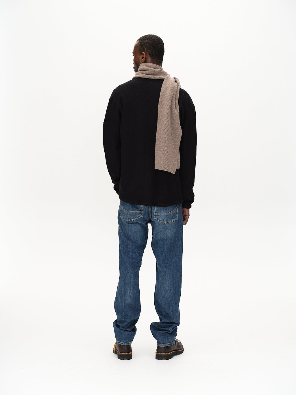 A man faces away from the camera wearing Gabbas Mattia Ls - Meteorite, with flatlock stitching, paired with light blue jeans, brown shoes, and a beige scarf over his shoulder against a plain white backdrop.