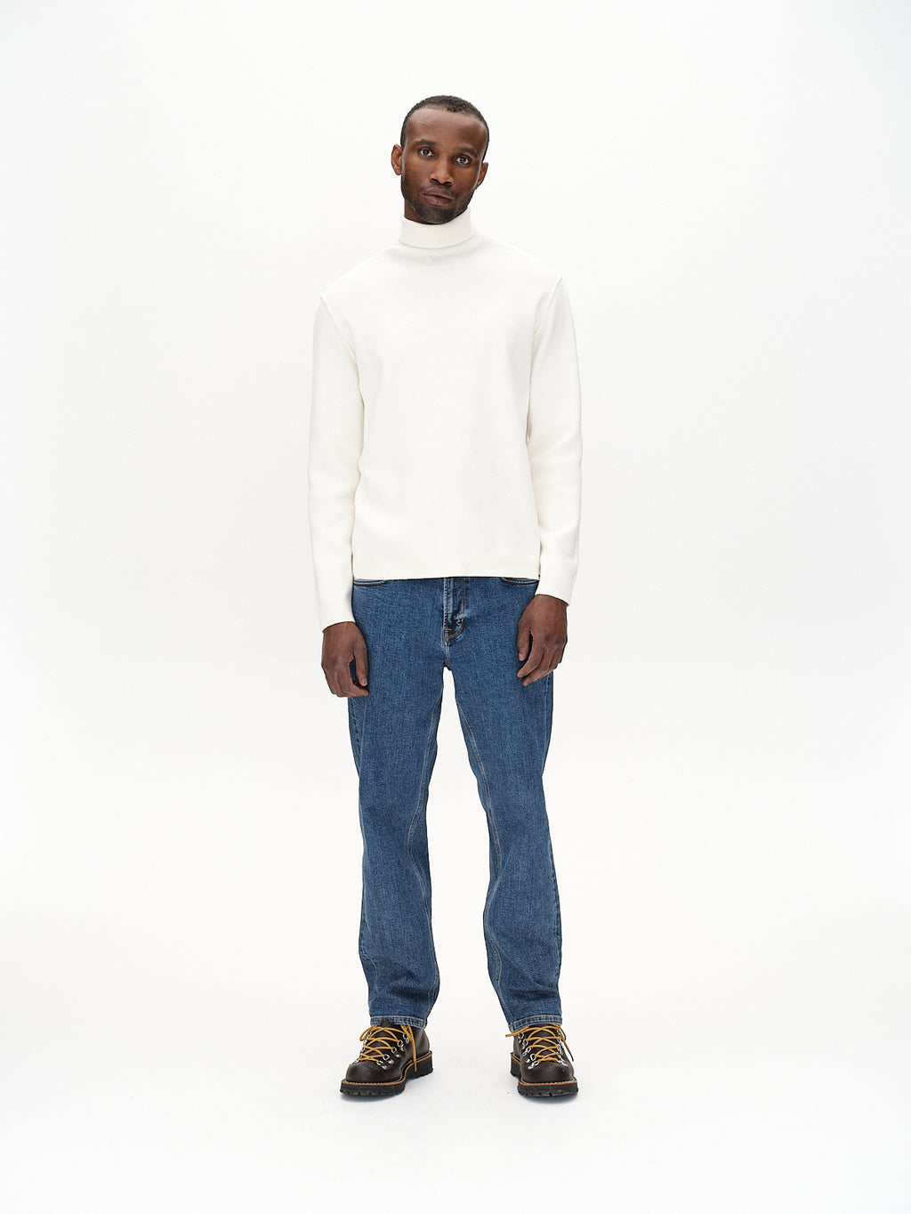 A man poses against a plain white backdrop in a Gabba Blanko High Ls - Off White turtleneck, blue jeans, and brown shoes with yellow laces. His hands rest by his sides, and he maintains a neutral expression.