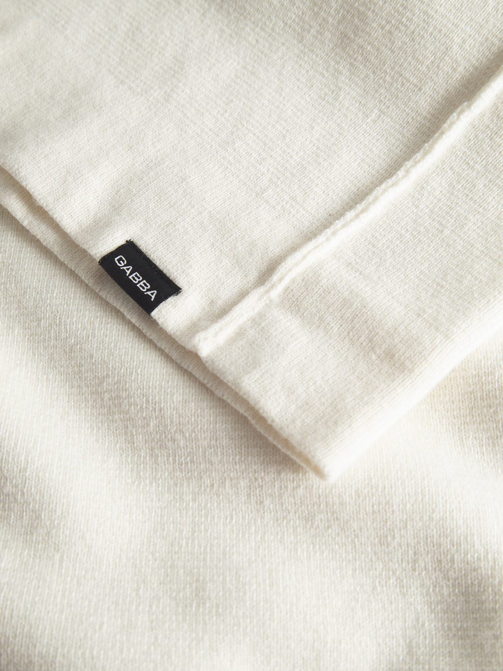 Close-up of a folded off-white Blanko High Ls sweater with a small black tag reading Gabba. The luxurious Milano knit fabric looks soft and textured, emphasizing craftsmanship and quality, making it a perfect addition to any stylish wardrobe.