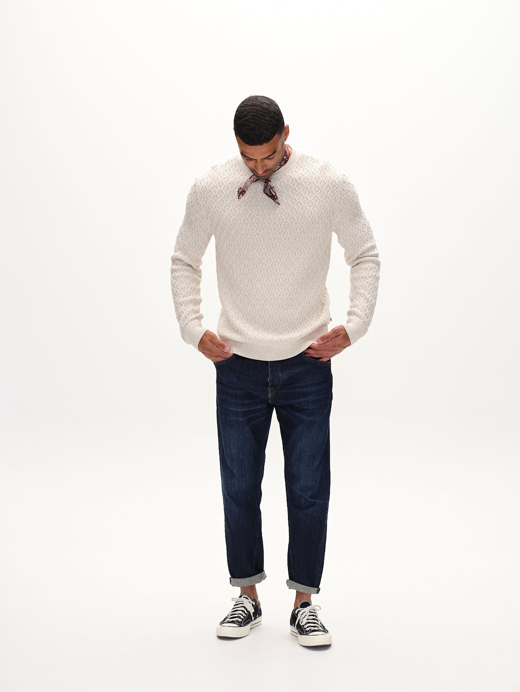 A person in the Gabba Aksla Ls - Egret, a cream sweater with a honeycomb knit, pairs it with dark blue jeans and black sneakers against a white backdrop. The 100% cotton sweater ensures comfort as they look down, hands by their sides, wearing a red bandana around their neck.