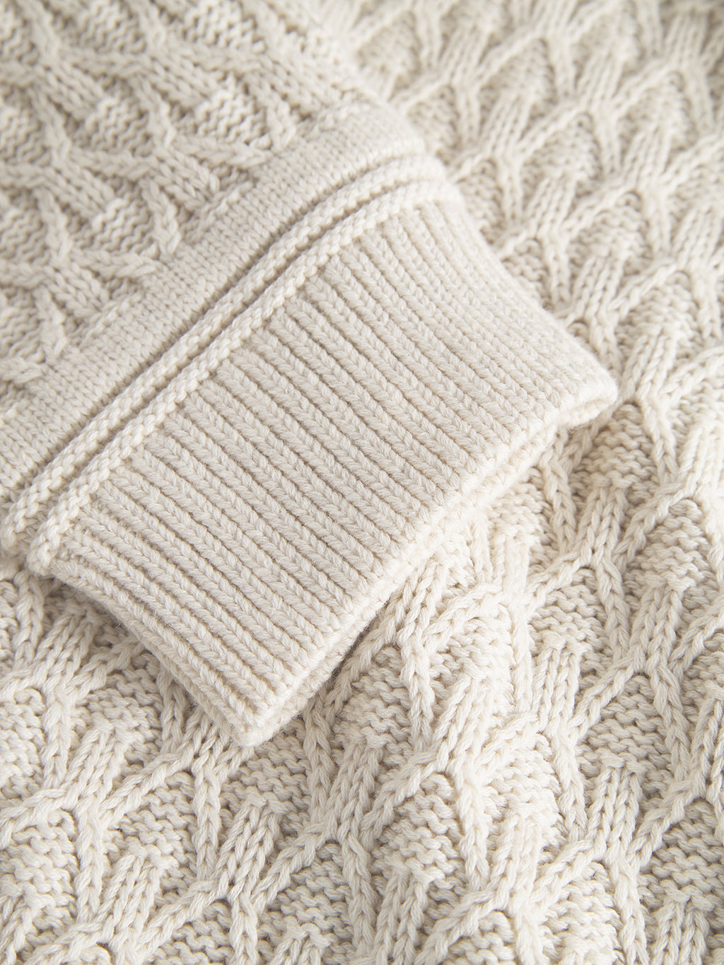 Close-up of Gabbas Aksla Ls - Egret, a cream-colored 100% cotton sweater featuring intricate cable patterns and ribbed cuffs. The honeycomb knit structure enhances comfort and highlights the knitworks texture and detail beautifully.