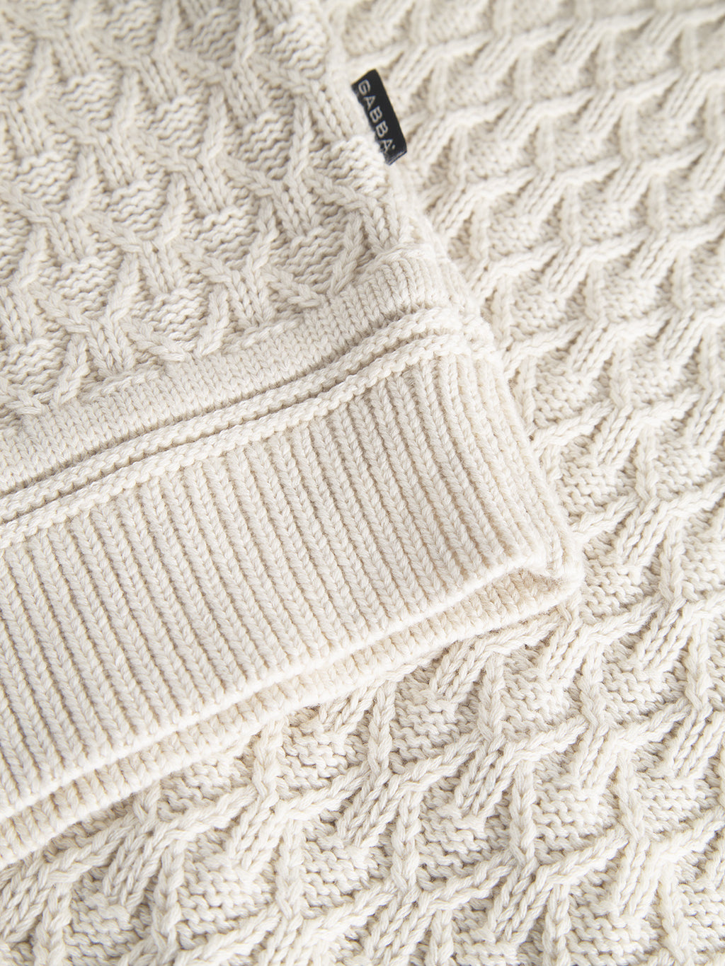 The Aksla Ls - Egret by Gabba is a cream-colored knit sweater featuring a detailed honeycomb pattern and ribbed cuffs. A small black label near the edge displays its 100% cotton composition for superior comfort.