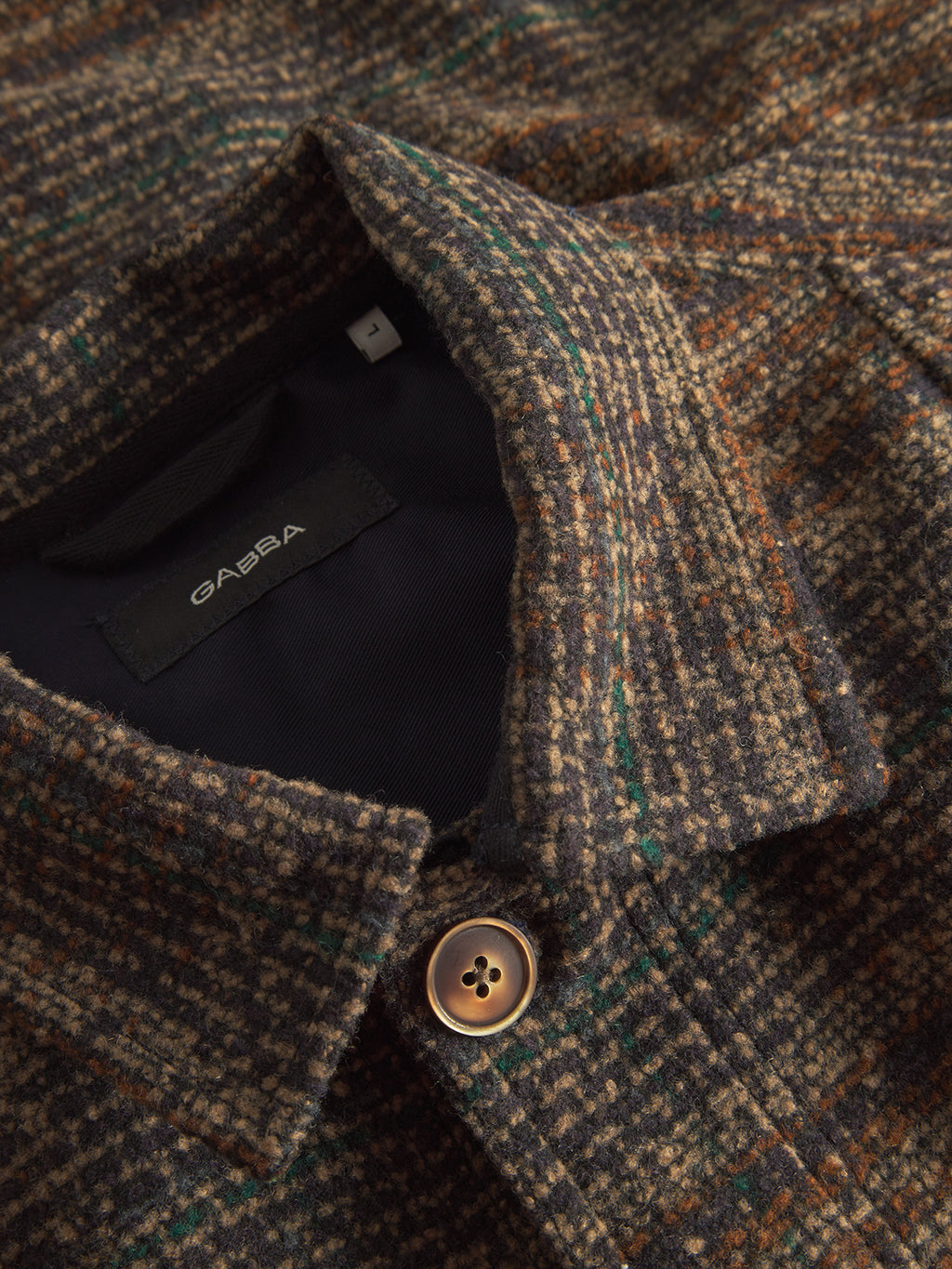 A close-up of the Oscar J Check wool blend jacket from Gabba shows its brown, green, and orange check pattern. It features a medium collar, a visible brown button, and an inside label with the brands name.