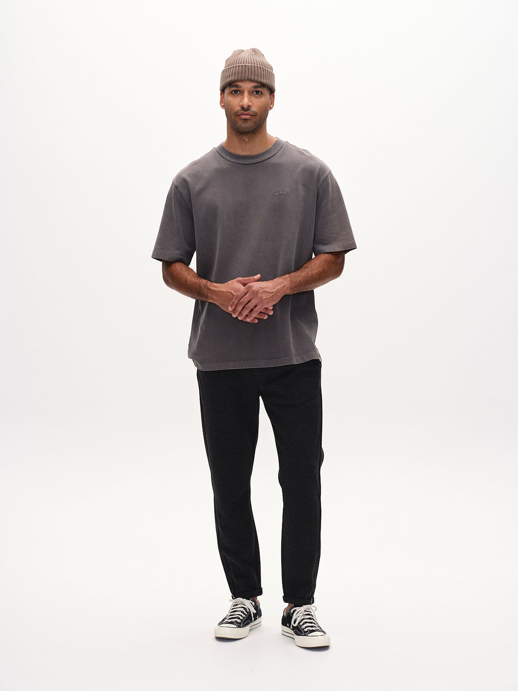 Against a white backdrop, a person wears Gabbas Pisa GKnit in Dk. Grey, black pants made from innovative structured fabric, a brown beanie, and black sneakers while clasping their hands.