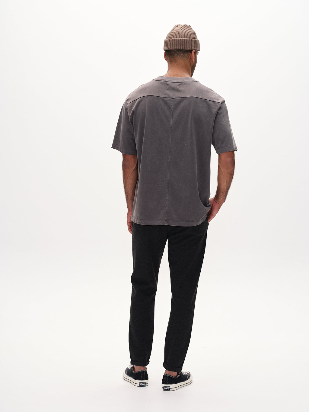 A person stands facing away, wearing a gray T-shirt, black pants crafted from innovative structure fabric, black shoes, and a Pisa GKnit in Dk. Grey by Gabba. The background is plain white.