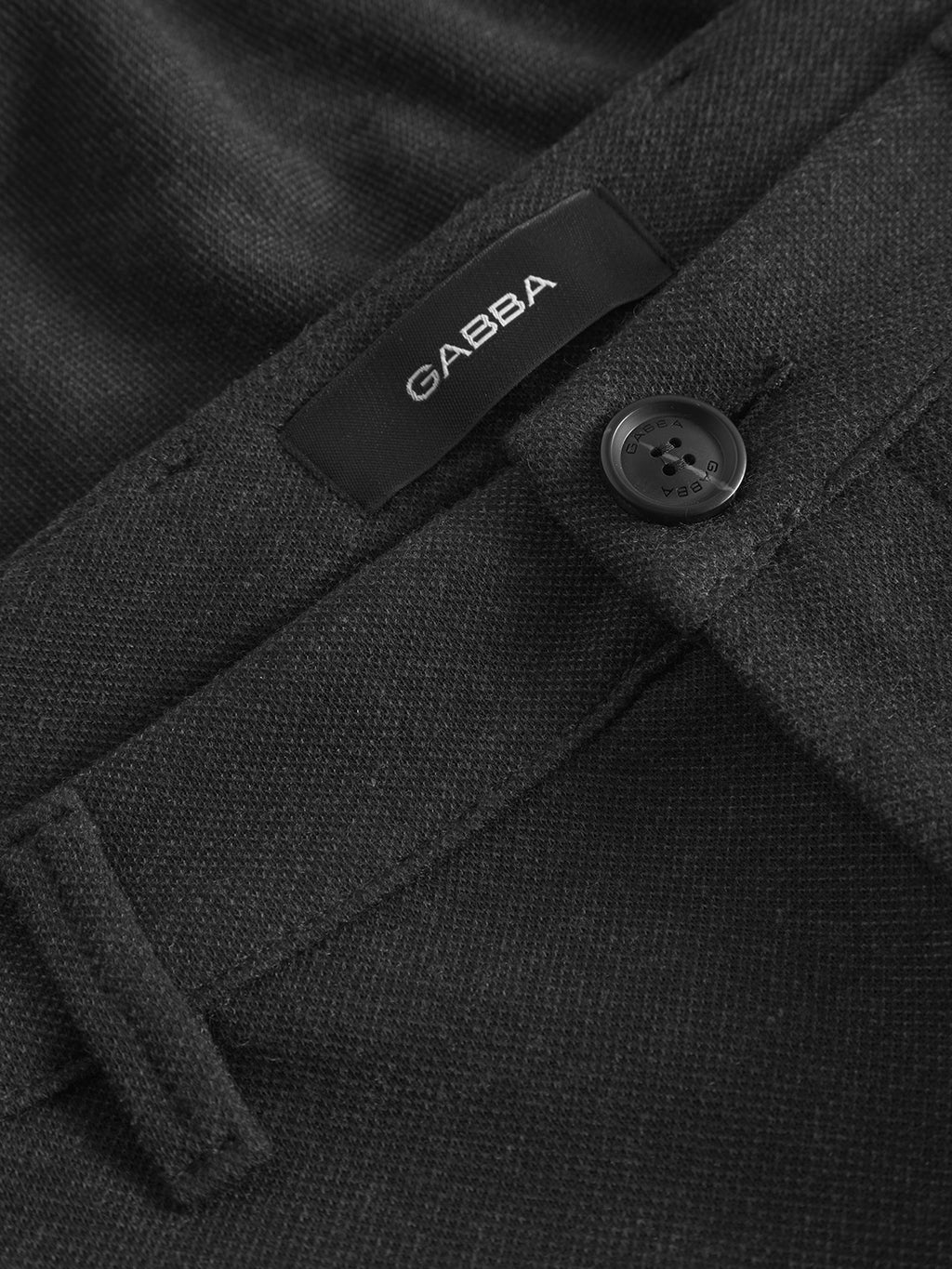 Close-up of Pisa GKnit - Dk. Grey pants by Gabba, featuring a button and belt loop. A black label with Gabba in white is sewn near the waistband, and the fabrics innovative structure offers a textured look.