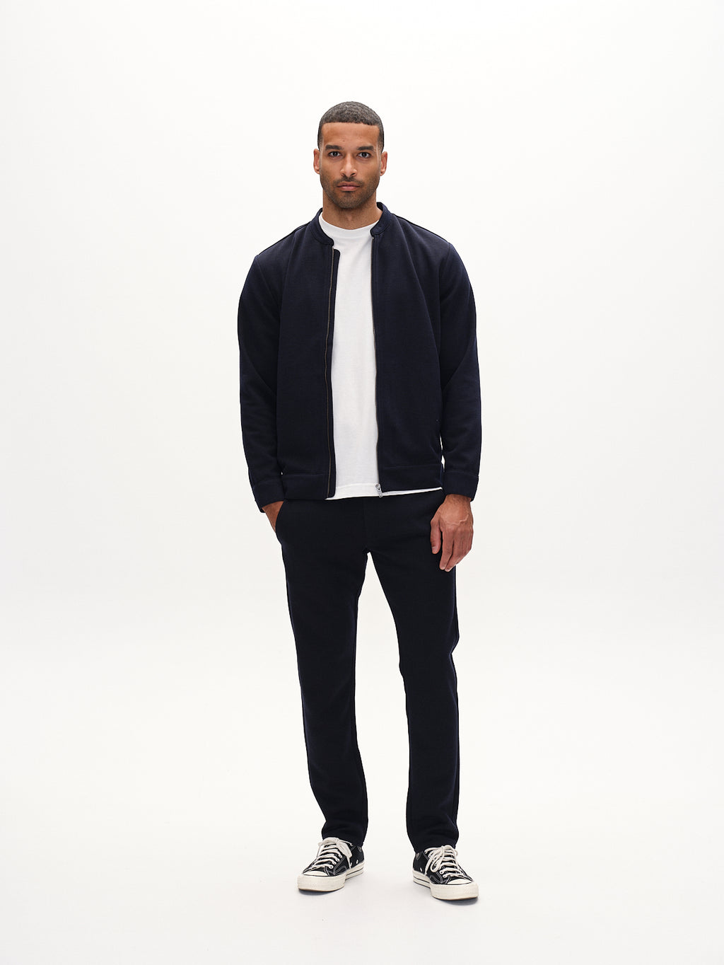 In a minimalistic setting, a person wears a black jacket and white shirt paired with Gabbas Paul GKnit pants in navy and black sneakers with white soles. They maintain a neutral expression against a plain white backdrop, exemplifying effortless everyday wear.