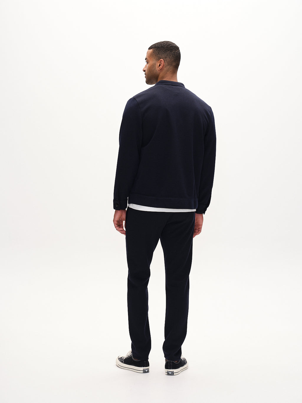 A man in a dark casual outfit showcases Gabbas Paul GKnit - Navy pants with a unique knitting technique against a plain white backdrop. Facing away in a jacket and sneakers, he slightly turns his head to the side, embodying everyday wear.