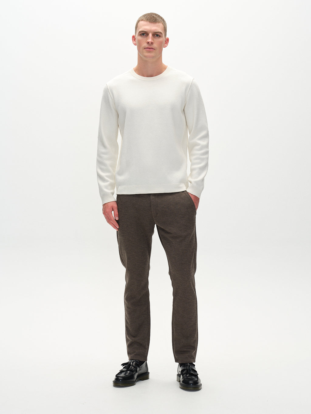 A person stands against a white background, wearing a white long-sleeve shirt and slim-fit Paul GKnit pants in Sand by Gabba, paired with black shoes. Their neutral expression and pocketed hands exude a sleek, composed vibe.