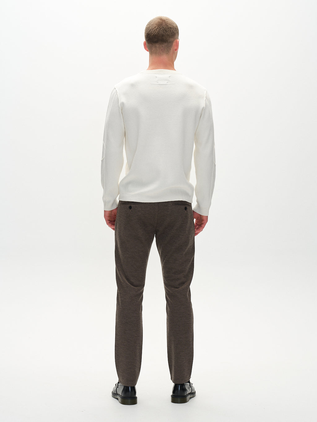 A person with short hair shows their back, wearing a white long-sleeve shirt, Gabbas Paul GKnit pants in sand color, and black shoes against a plain white background.