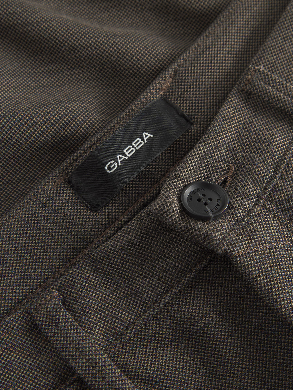 A close-up of a textured brown garment with a black button and GABBA label. Made from structured fabric with a subtle checkered pattern, it features visible stitching indicative of high-quality craftsmanship. This slim-fit piece highlights the meticulous design seen in the Paul GKnit - Sand by Gabba.