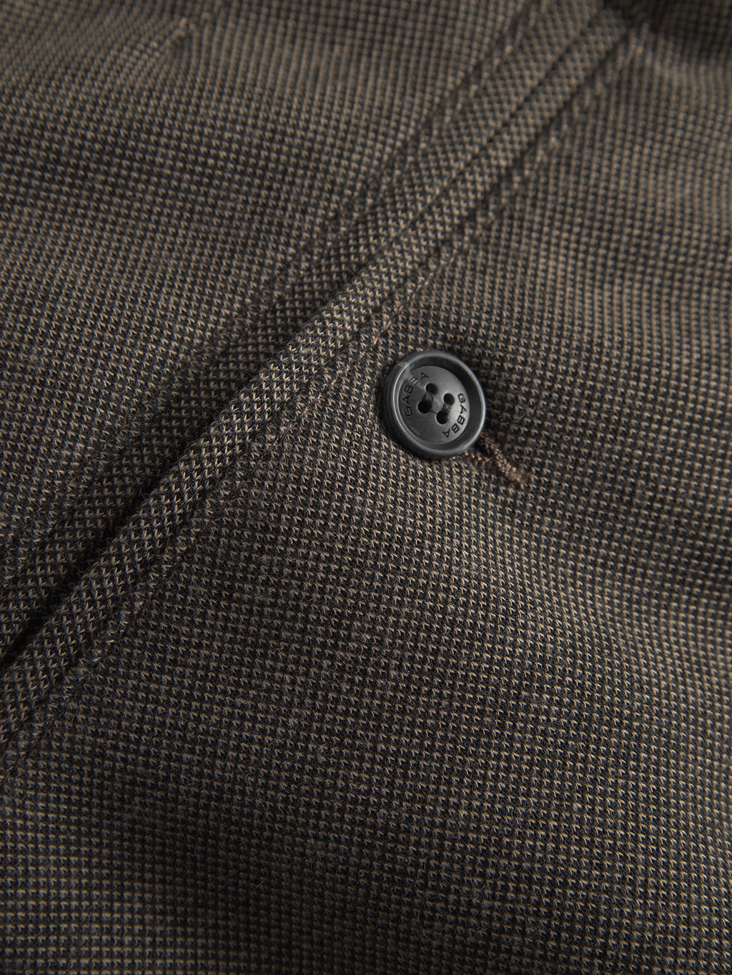 Close-up of a textured, structured fabric in dark brown and black checkered pattern with a visible seam. A black button with four holes is sewn on near the seam, showcasing detailed stitching reminiscent of Paul GKnit by Gabba.