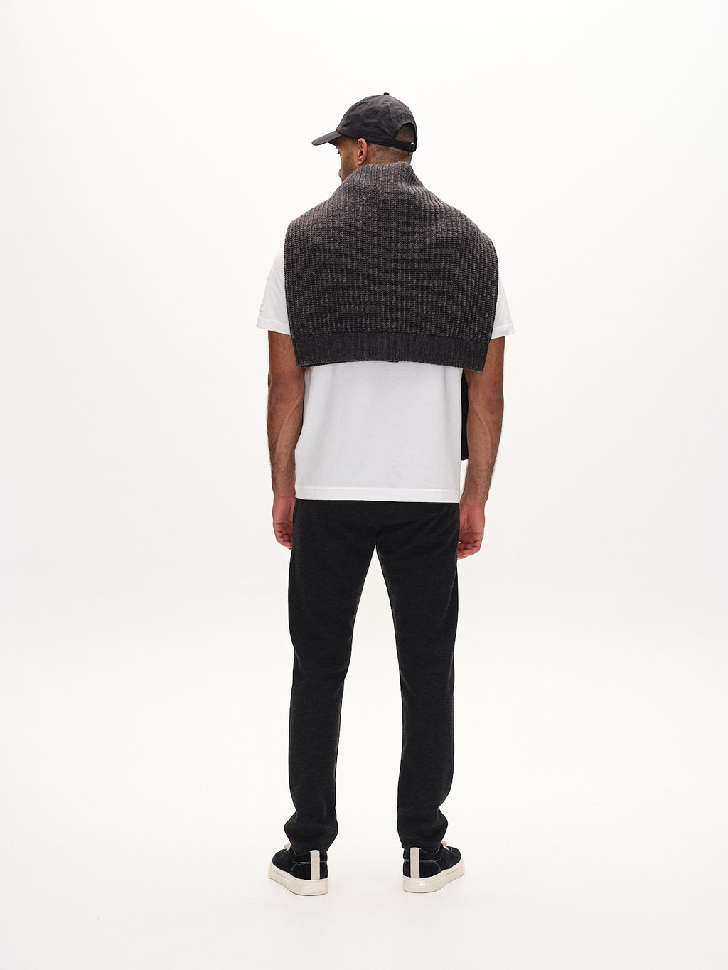 A man stands with his back to the camera, wearing a white t-shirt, slim-fit dark joggers, sneakers, and a black cap. Draped over his shoulders is the Gabba Paul GKnit sweater in Dk. Grey. The ensemble embodies everyday style against a plain white backdrop.