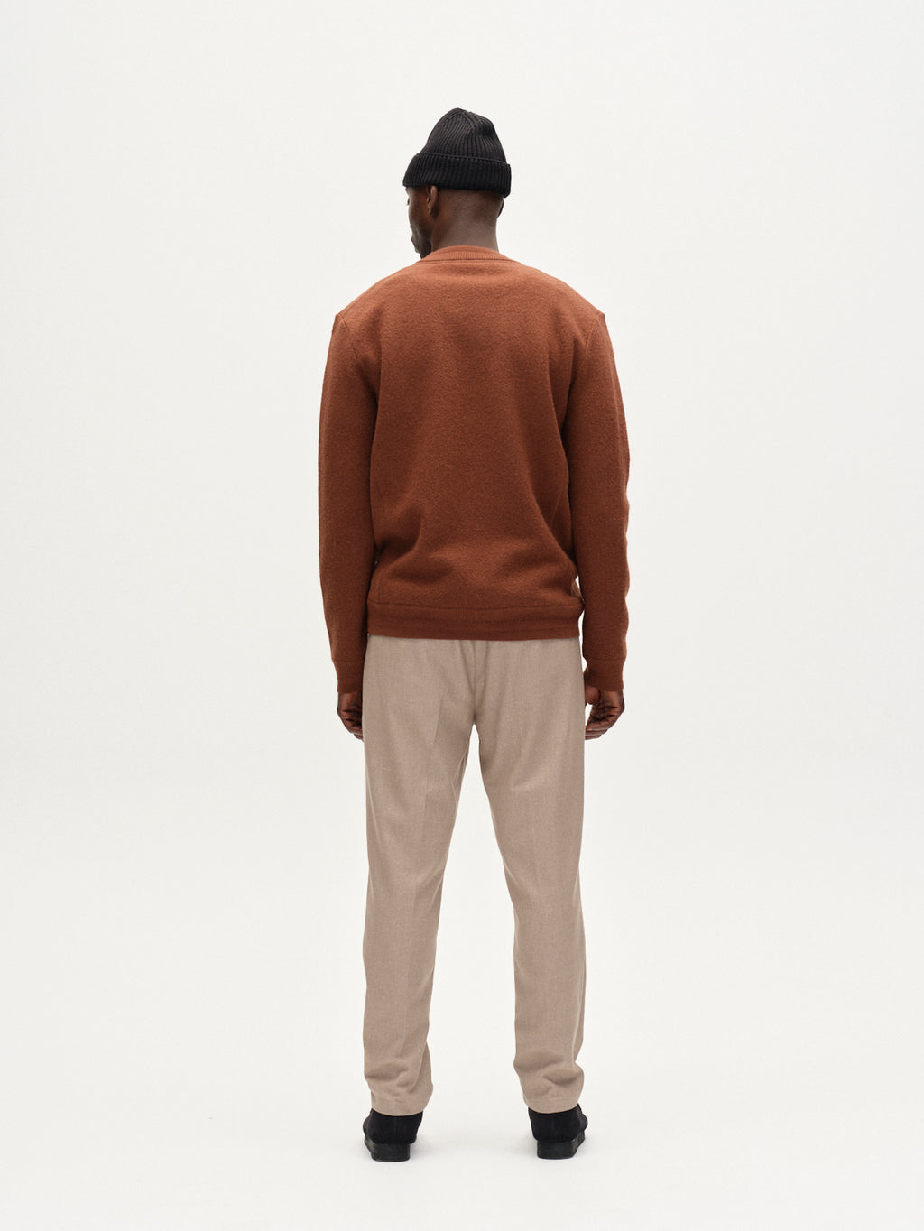 A man stands with his back to the camera, wearing Gabbas Monza Shafi - Brown sweater, a black beanie, beige relaxed straight fit brushed cotton pants, and black shoes against a plain white background.