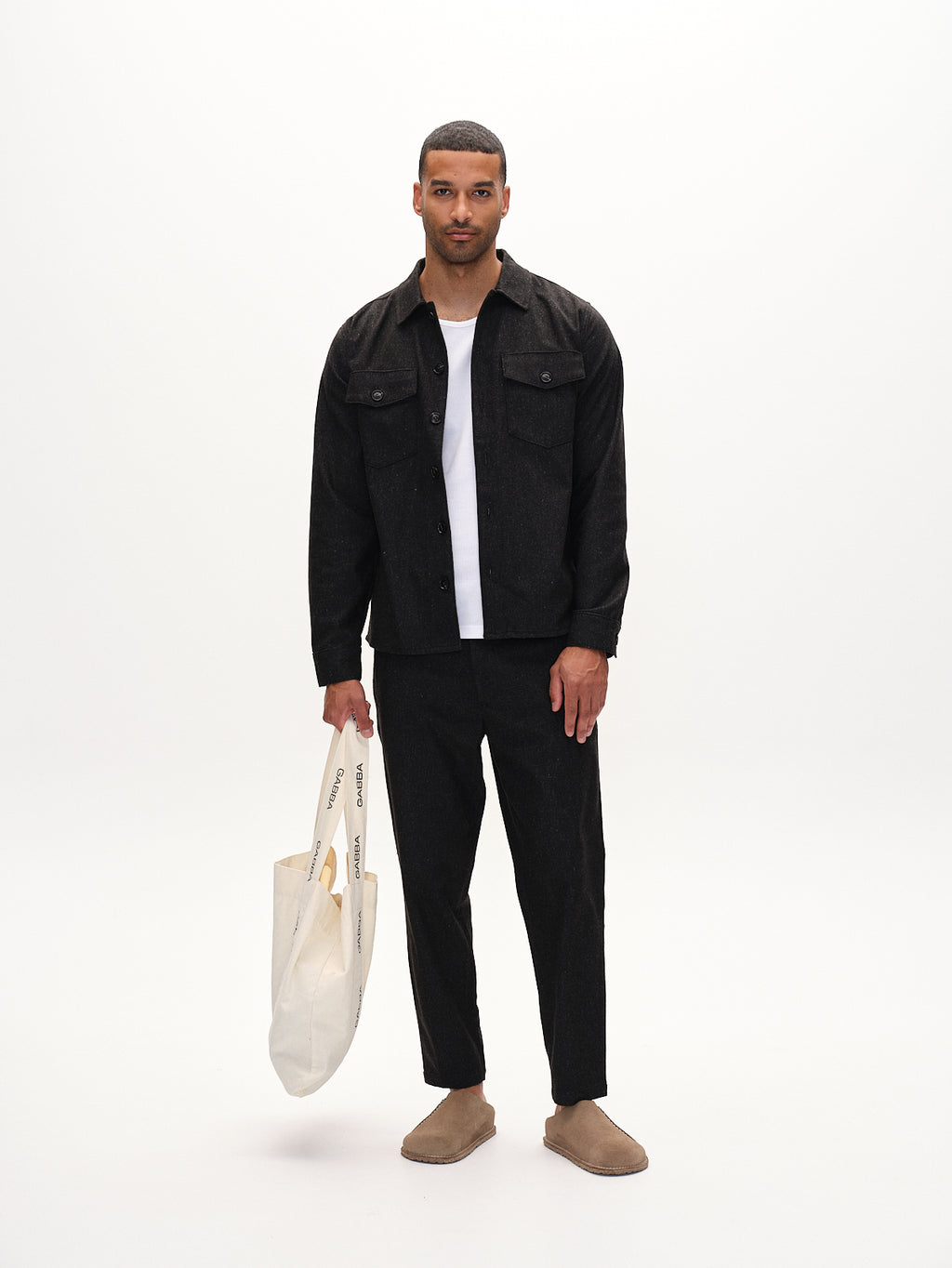 A man stands against a white background wearing Gabbas Monza Shafi - Black jacket, a white t-shirt, brushed stretch trousers, brown slip-on shoes, and holding a cream tote bag—ideal for modern dressing.