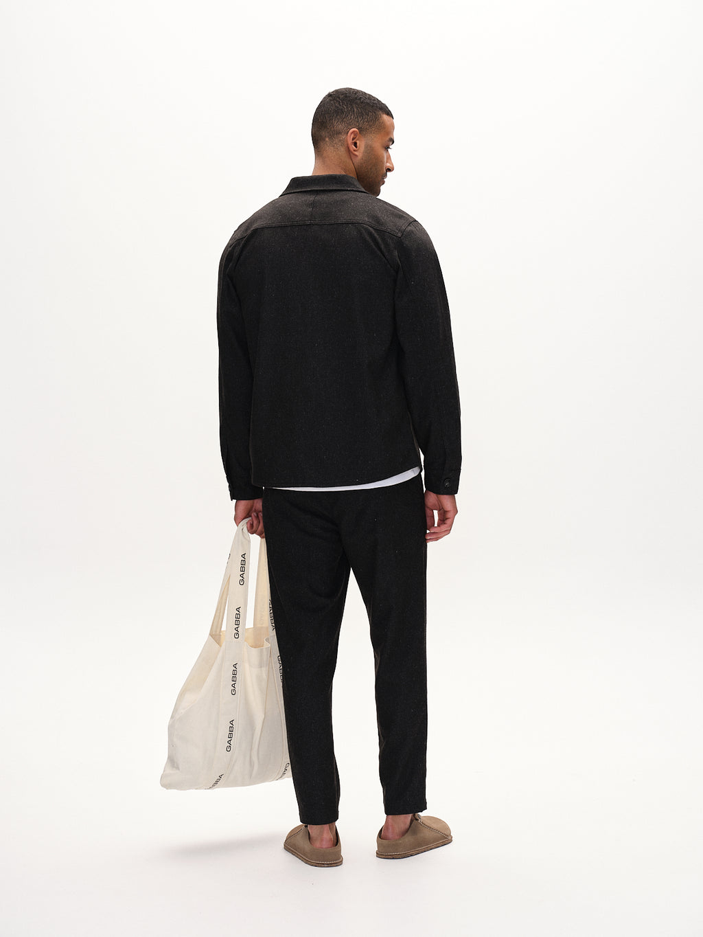 A man faces away, wearing Gabbas Monza Shafi - Black jacket and brushed stretch trousers with brown slippers. He holds a large white canvas tote bag featuring black text against a plain white background.