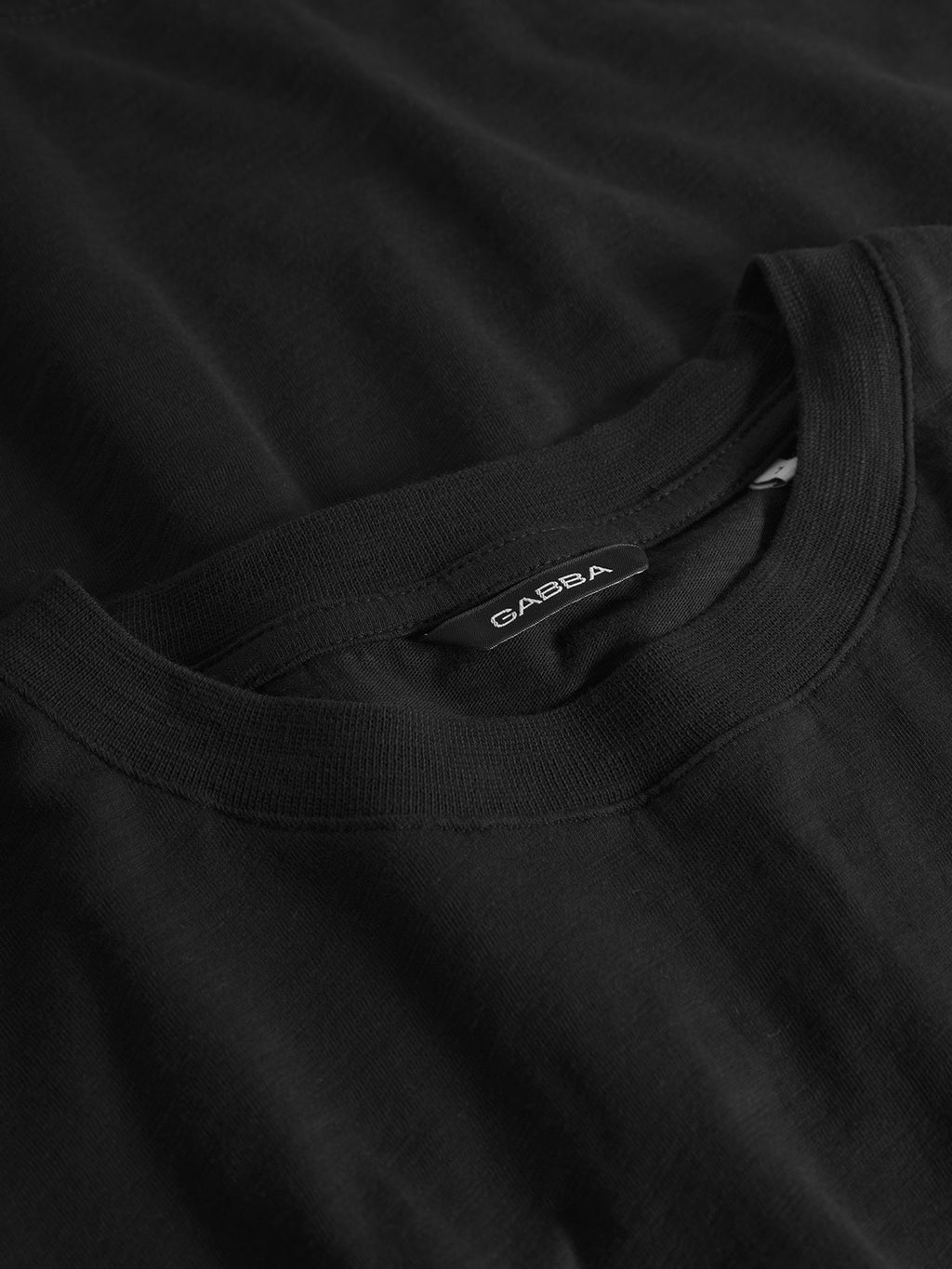 A close-up of a neatly folded black T-shirt reveals a visible label inside the collar displaying Gabba in white text. This Pure Ss - Meteorite tee is crafted from midweight slub jersey, offering a smooth yet slightly textured fabric for a regular fit look.