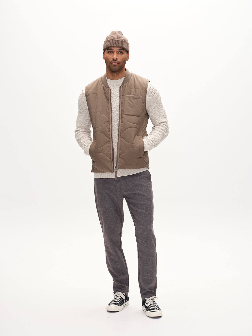 A man stands against a white backdrop wearing a beige beanie and quilted vest over a cream sweater. His hands rest in his pockets, complementing his black sneakers, while Gabbas Pisa Field - Grey pants crafted from high-stretch cotton-blend enhance his metro style as he faces forward.