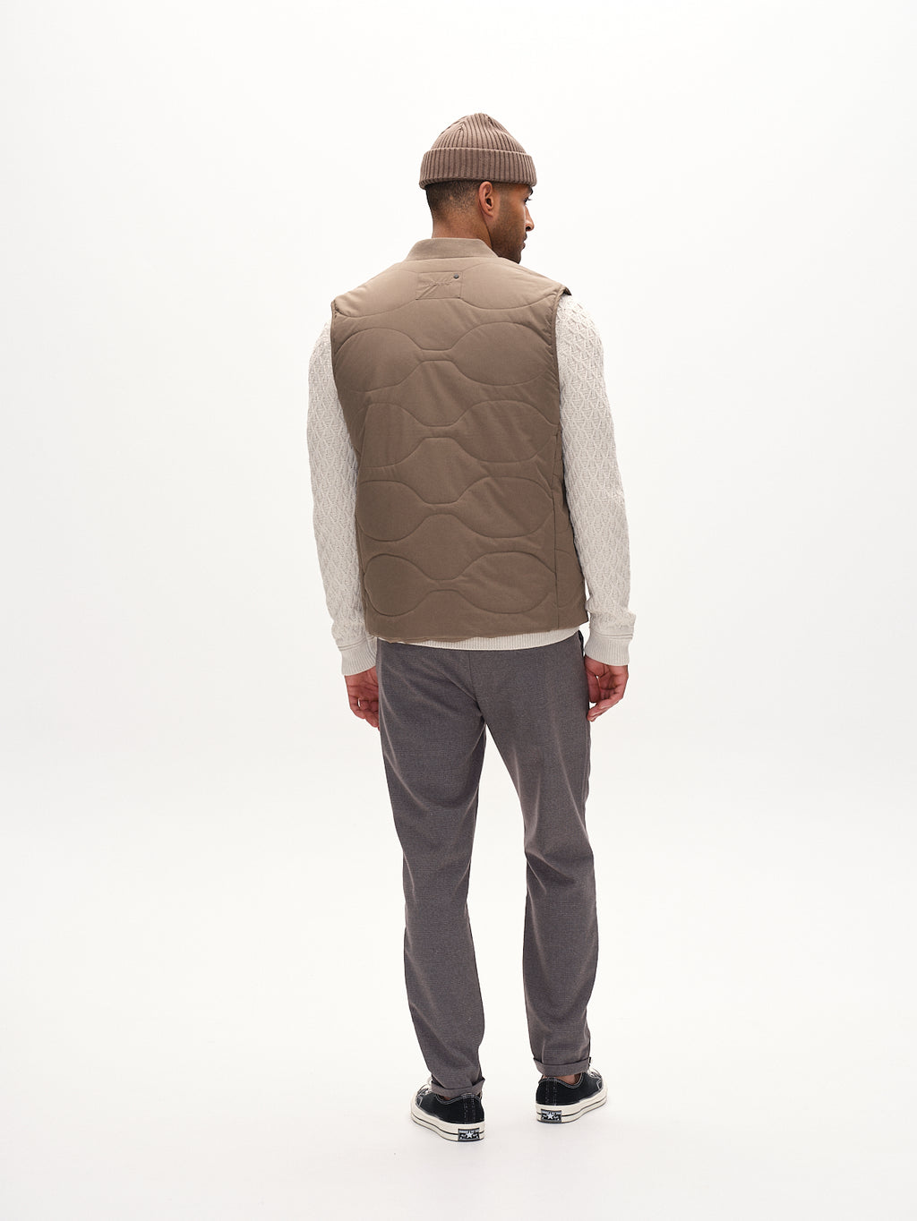 A man in a beige beanie, quilted vest, white sweater, and gray pants stands against a plain white background wearing black and white sneakers. His metro style features Gabbas Pisa Field - Grey zippers for subtle accents.