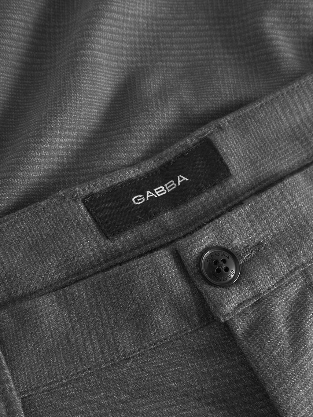 Close-up of gray textured high-stretch cotton-blend fabric with a black Gabba label. A detailed button is near the edge, and the material features Gabba Pisa zippers, indicating its part of a Pisa Field - Grey metro style clothing item like pants or a jacket.