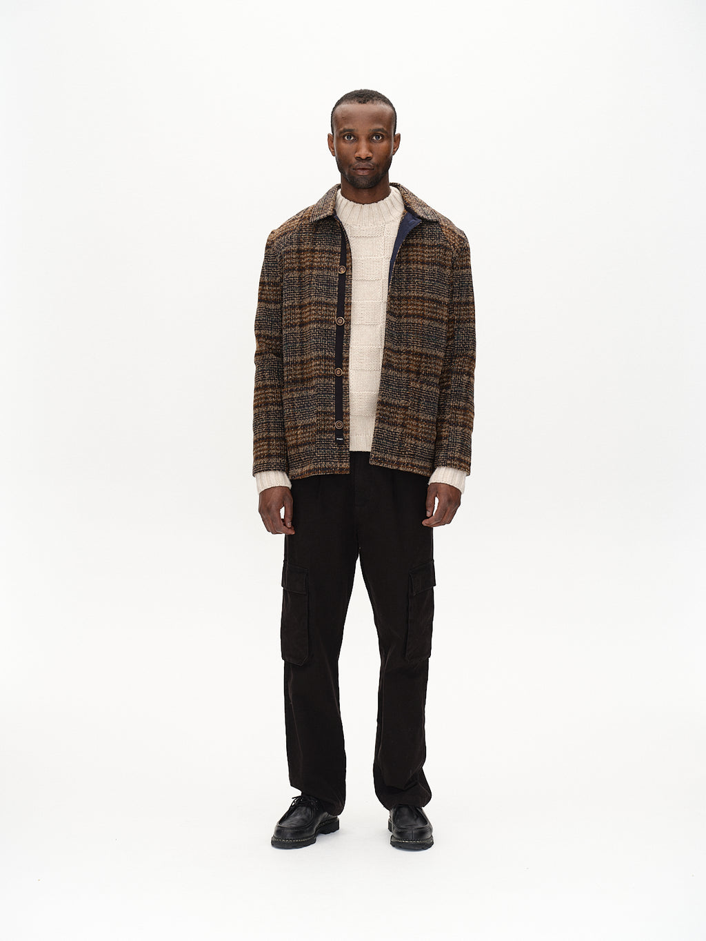 A person in a brown plaid jacket and light sweater stands confidently against a white background, wearing Gabbas Verona Ent Poc - Meteorite dark cargo pants with spacious side pockets and black shoes, looking directly at the camera.