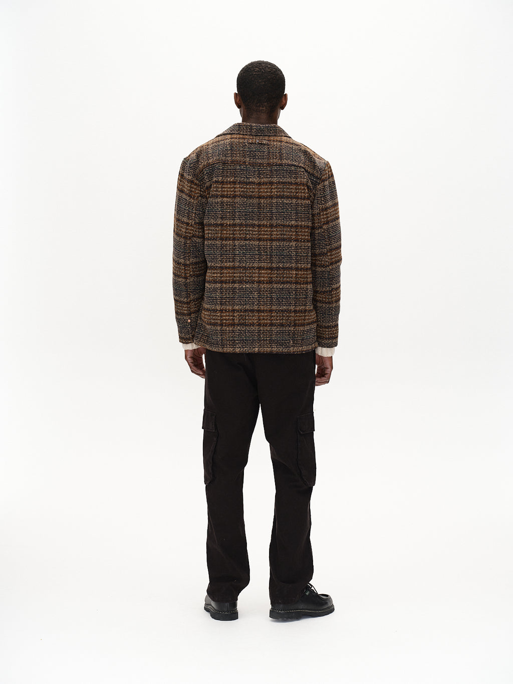 Facing away from the camera, a person wears a Verona Ent Poc - Meteorite by Gabba, featuring spacious side pockets and a brown plaid jacket. A white backdrop emphasizes the garment dye finish of their outfit in this minimalistic setting.
