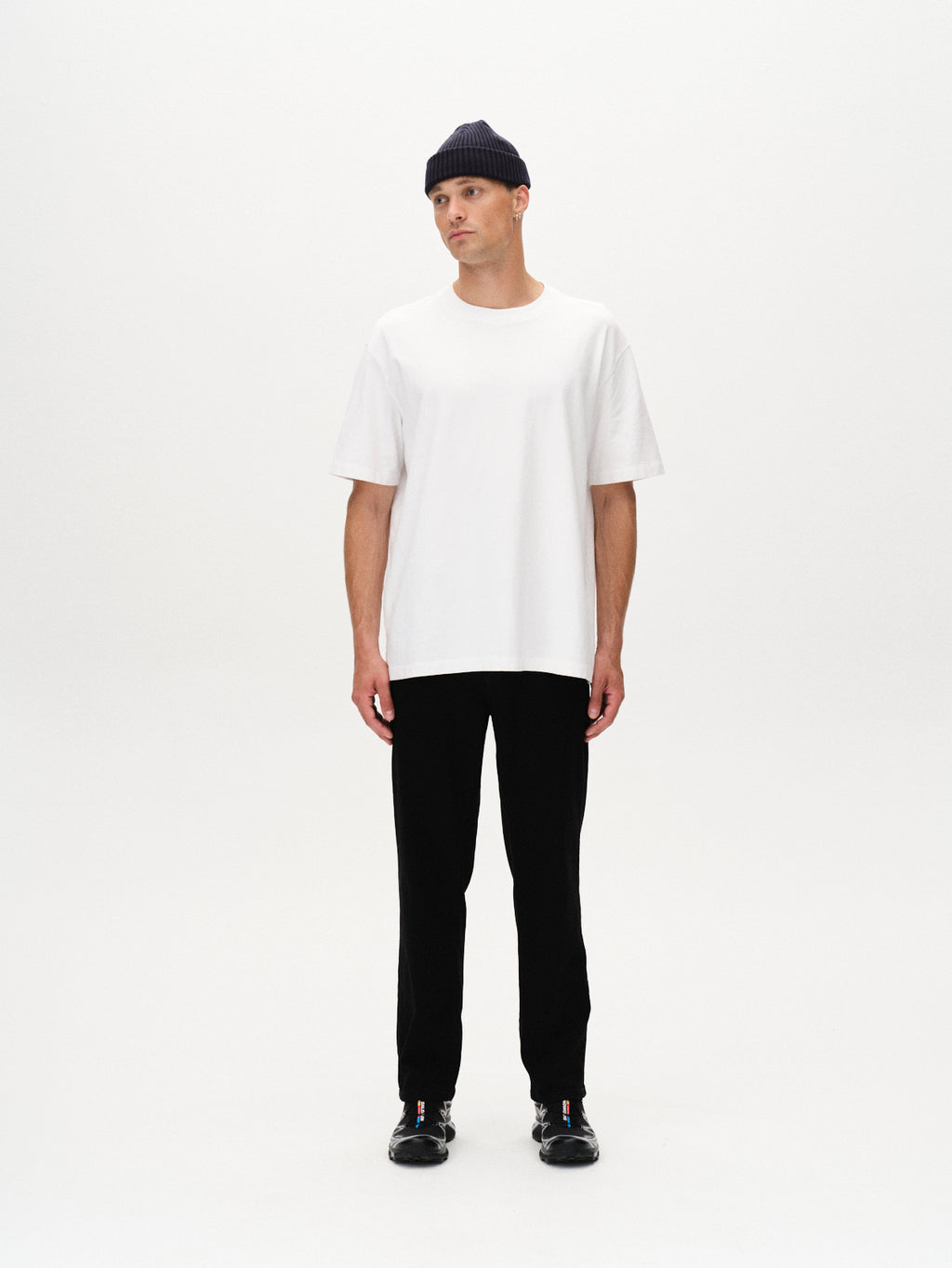 A person stands against a plain white background, wearing a black knit beanie, black pants, black shoes, and the Gabba Nigel Wix Boxy Hour SS t-shirt in white. They face forward with a neutral expression and arms at their sides.