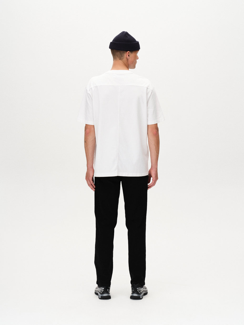 A man faces away from the camera wearing the Gabba Nigel Wix Boxy Hour SS white t-shirt, paired with black pants, black sneakers, and a black beanie, set against a plain white background.