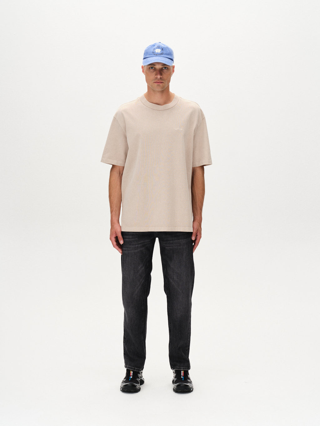 A man stands facing forward against a plain white background, wearing the Gabba Nigel Wix Boxy Hour SS t-shirt in Sand Cashmere, paired with dark jeans, black shoes, and a light blue baseball cap.