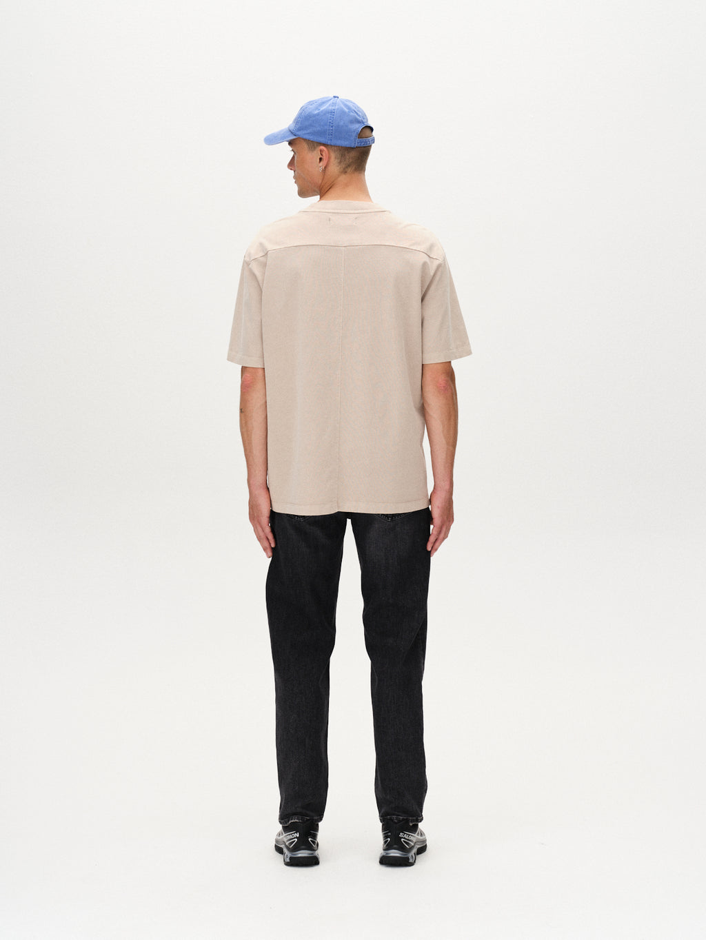 A man faces away from the camera, wearing the Gabba Nigel Wix Boxy Hour SS - Sand Cashmere t-shirt with black pants, black sneakers, and a light blue baseball cap against a plain white background.