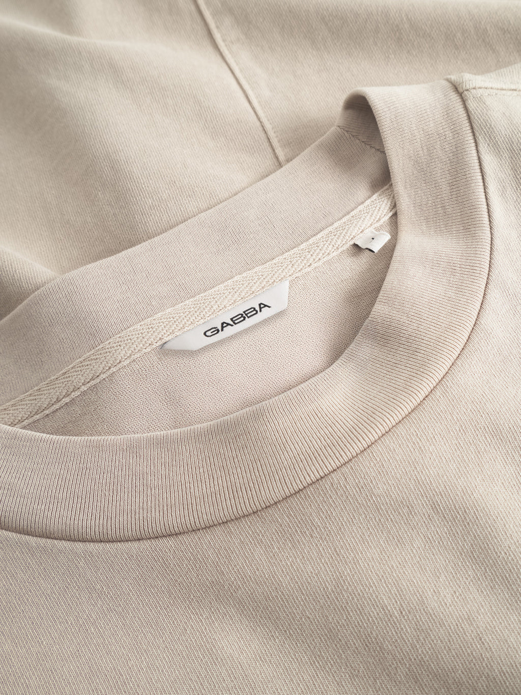 A close-up of the Gabba Nigel Wix Boxy Hour SS sweatshirt in sand cashmere, featuring a white Gabba tag, lying atop another modern-fit casual t-shirt in similar tones.