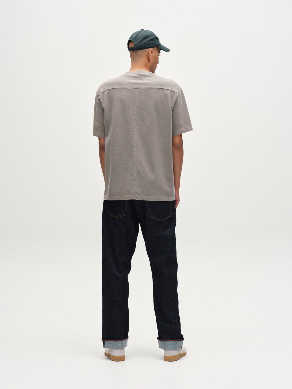 A person wears the Gabba Nigel Wix Boxy Hour SS in Dust Sage Green, paired with dark jeans and a green baseball cap, standing with their back to the camera against a plain white background.