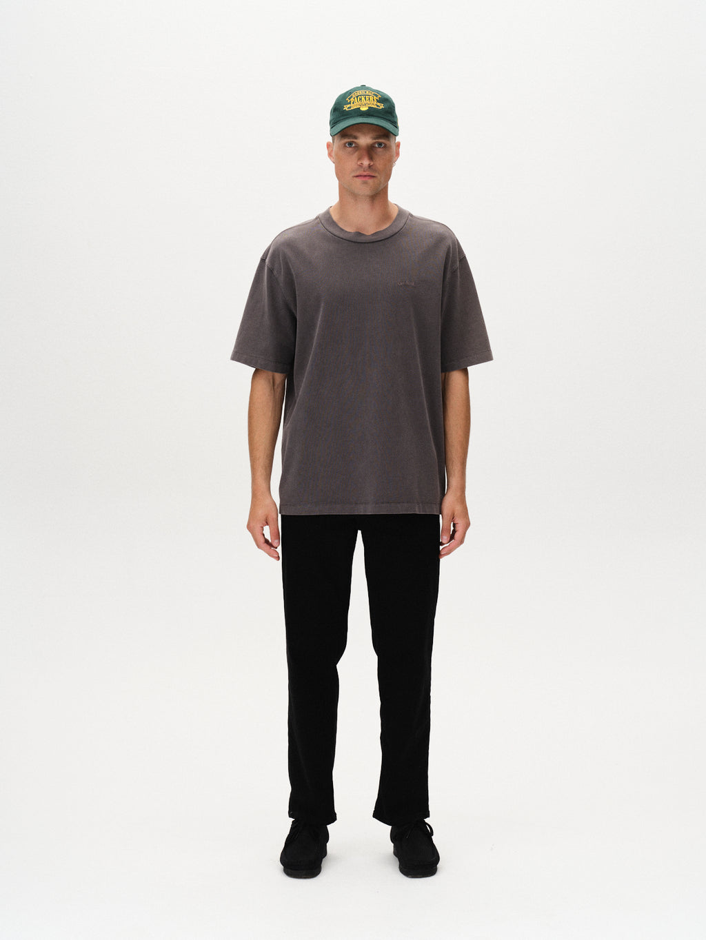 A person stands facing forward against a plain white background, wearing a dark green cap, the Gabba Nigel Wix Boxy Hour SS tee in Dk. Grey, black pants, and black shoes.