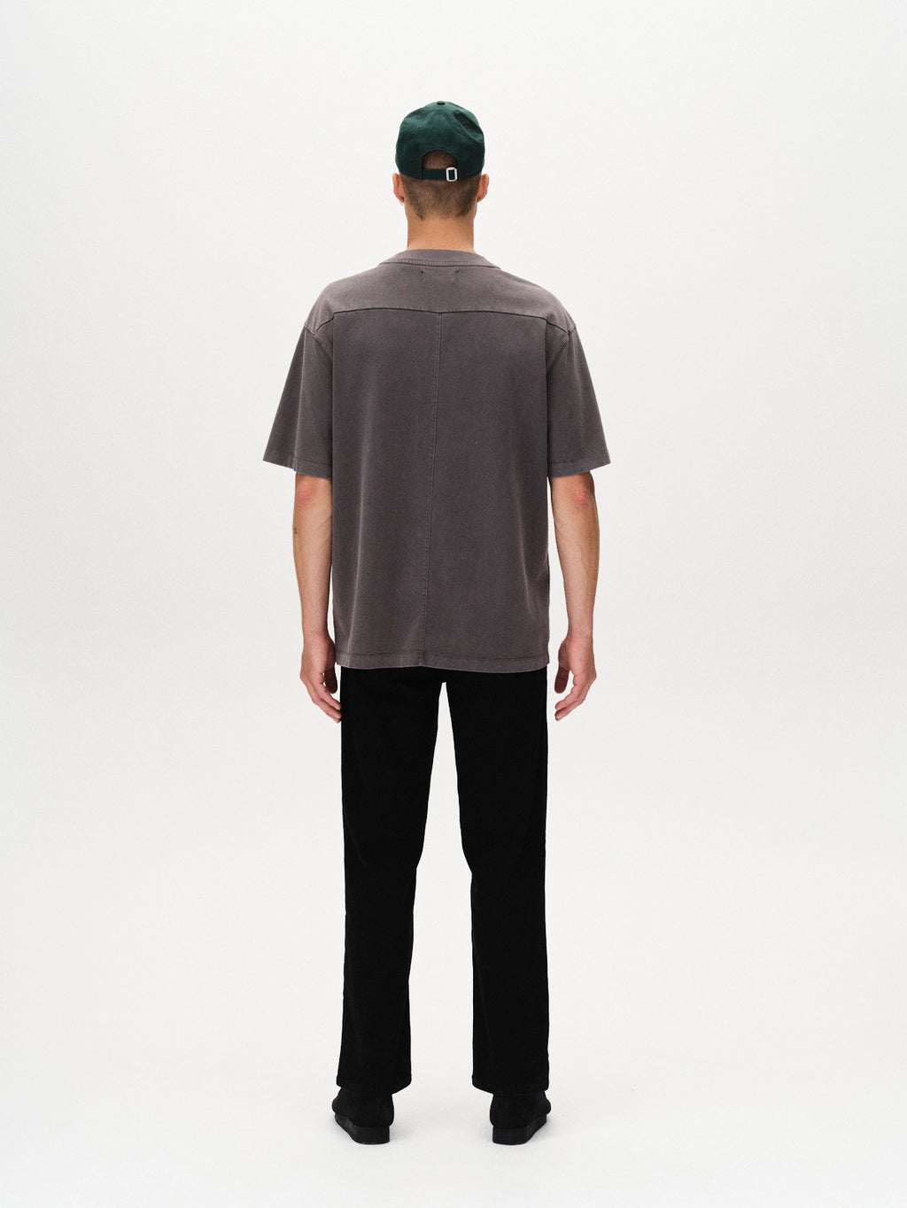 A person stands facing away from the camera, wearing a dark green cap, black pants, black shoes, and a Gabba Nigel Wix Boxy Hour SS tee in Dk. Grey with short sleeves against a plain white background.