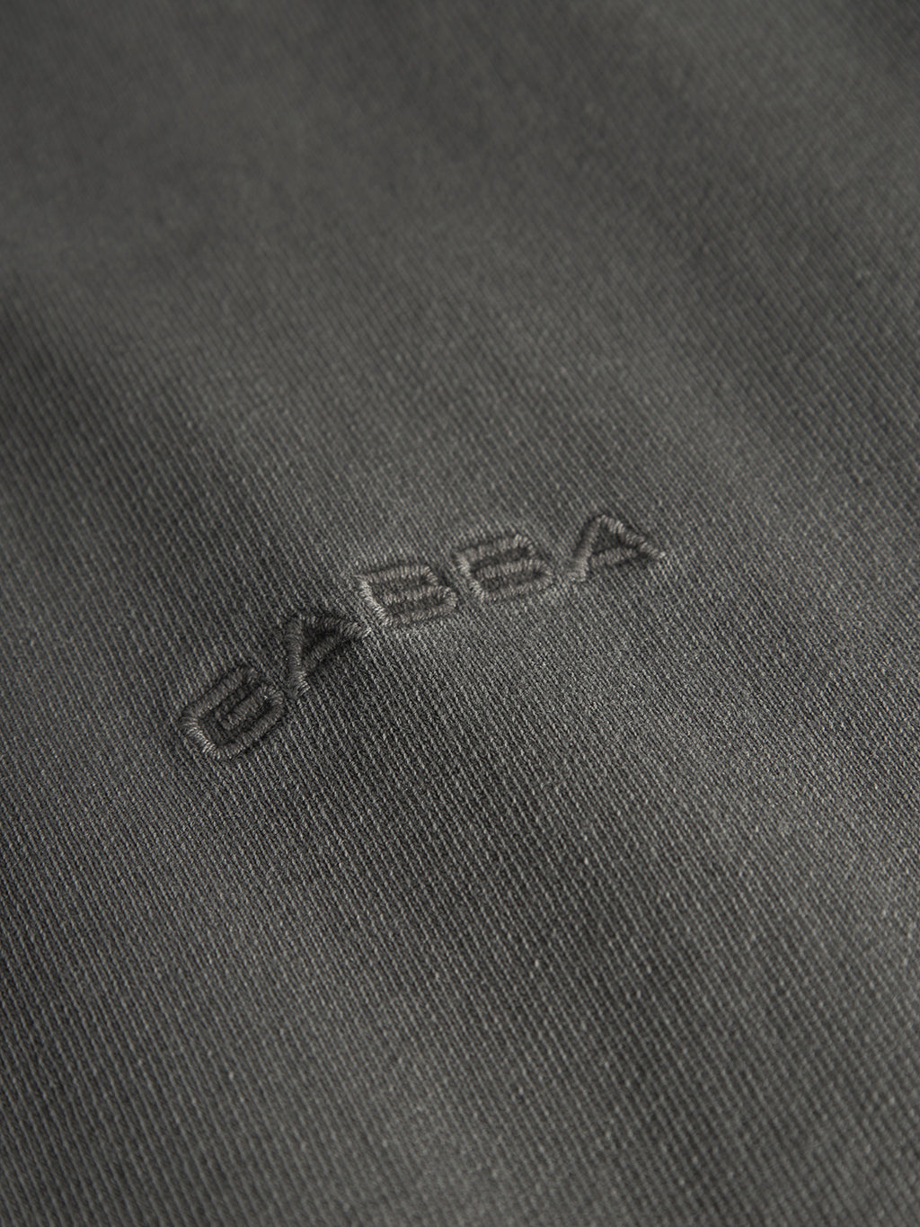 Close-up of a dark grey fabric with Gabba embroidered in the center. The texture is slightly ribbed, similar to a relaxed fit tee, adding subtle visual depth to the Nigel Wix Boxy Hour SS by Gabba.