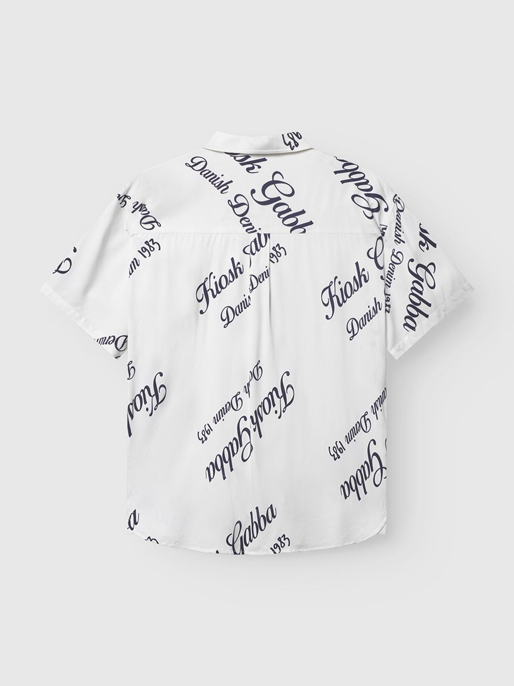 The Gabba Jens Over Resort Kiosk SS in Navy Pattern is a short-sleeve, collared shirt made from 100% viscose, featuring black cursive and serif text such as “Kiosk,” “Gabba,” “Danish,” and “Founded 1983.” Shirt shown flat with back facing up.