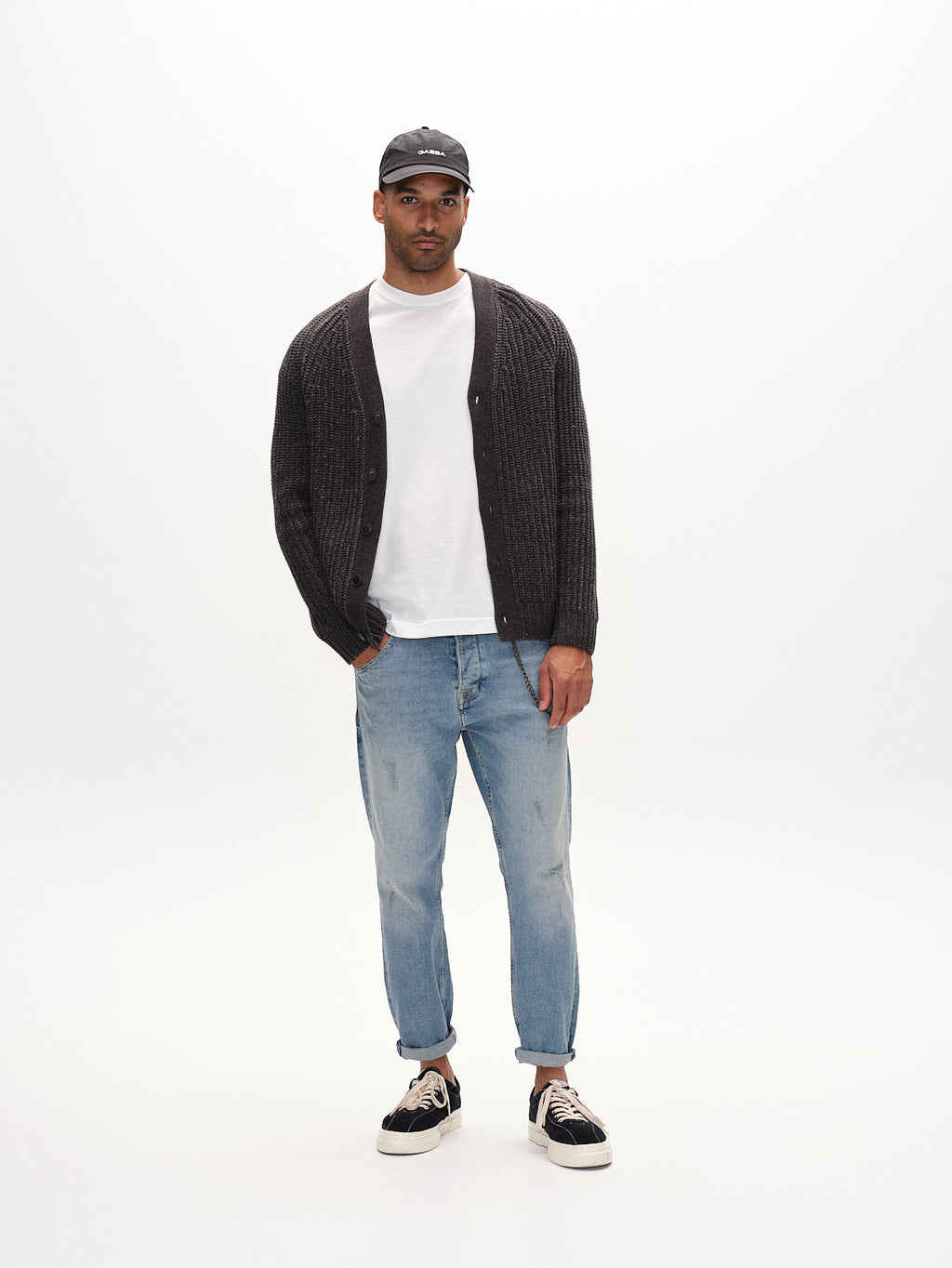 A man in a black cap, white t-shirt, dark gray cardigan, Gabbas Alex Cros light blue denim with a relaxed fit made from recycled cotton, and black sneakers stands on a plain white background. His hands are in his pockets, and the jeans are slightly rolled at the cuffs.
