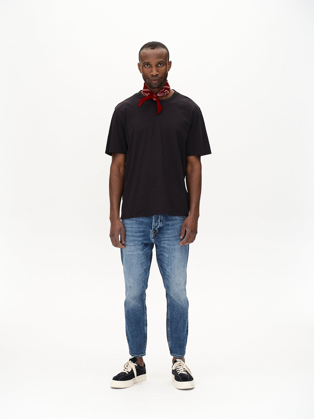 A person stands against a plain white background wearing a black t-shirt and Gabbas Alex Cros Mid Blue Denim in a relaxed fit. Black sneakers with white laces and a red bandana around the neck complete the casual look.