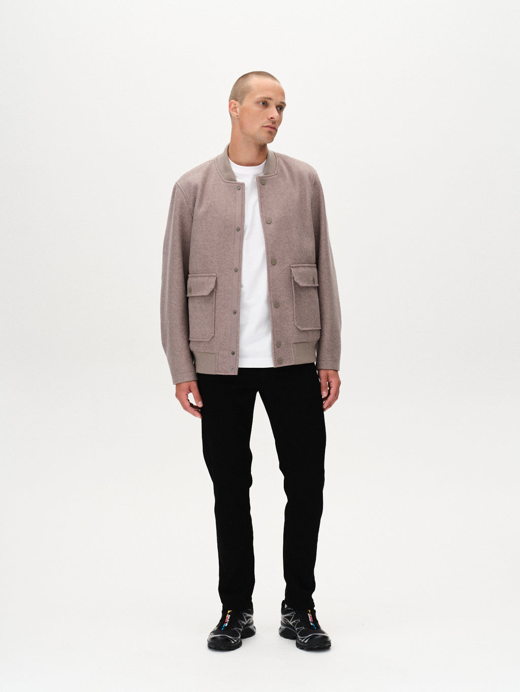 A man with short hair stands against a white background, wearing Gabbas Marc Sword - Black Denim pants, a slim fit light mauve jacket, white t-shirt, and black sneakers. He gazes slightly aside with a neutral expression—elegance meets edge.