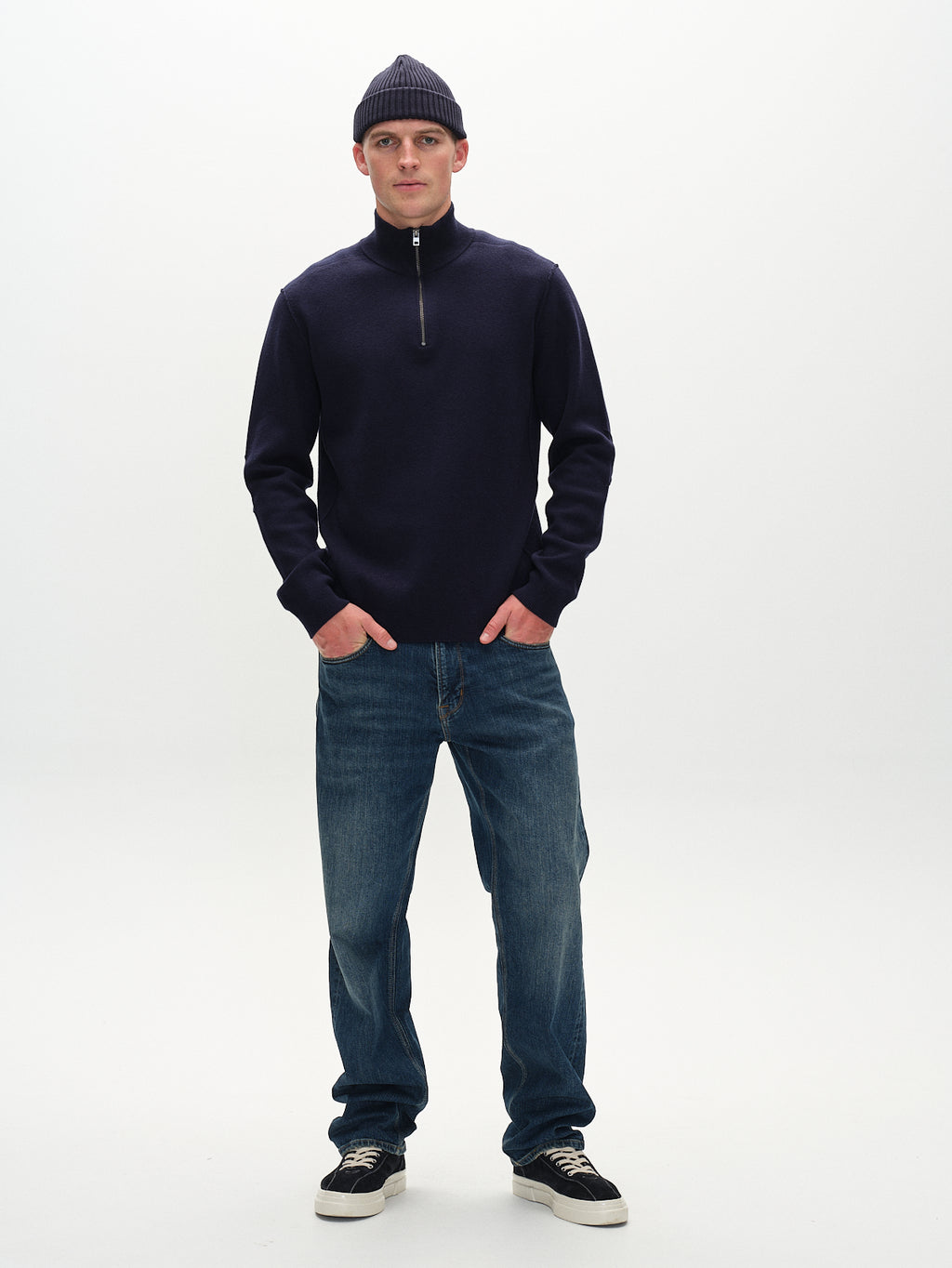 A man exudes modern elegance against a plain background, wearing Gabbas Max Loom Dark Blue Denim and a navy quarter-zip sweater. His dark beanie and black sneakers complement the outfit as he faces the camera with his hands in his pockets.
