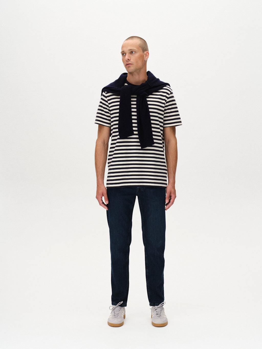 A man stands against a white background, wearing black and white striped tee, Marc Mego - Dark Blue Denim jeans by Gabba, white sneakers, and a black sweater over his shoulders. He gazes slightly to the side.